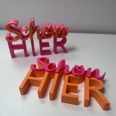 Zweifarbiger 3D-Schriftzug „Schön hier" in Orange und Hot Pink auf dem Sideboard