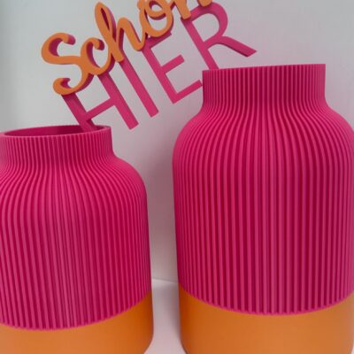 Wohndeko-Set aus der mittleren und großen geriffelten Vase und 3D-Schriftzug „Schön hier" in Orange und Hot Pink auf einem Sideboard